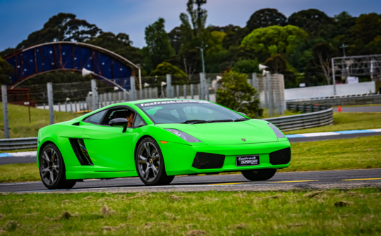 Bright green Lamborghini sports car on a race track with grandstands and trees in the background, captured in a front-three-quarter view.