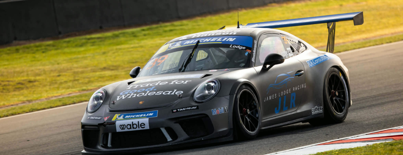 Gray Porsche race car with a large rear wing on a racetrack, sponsor decals visible along the sides and hood edges.