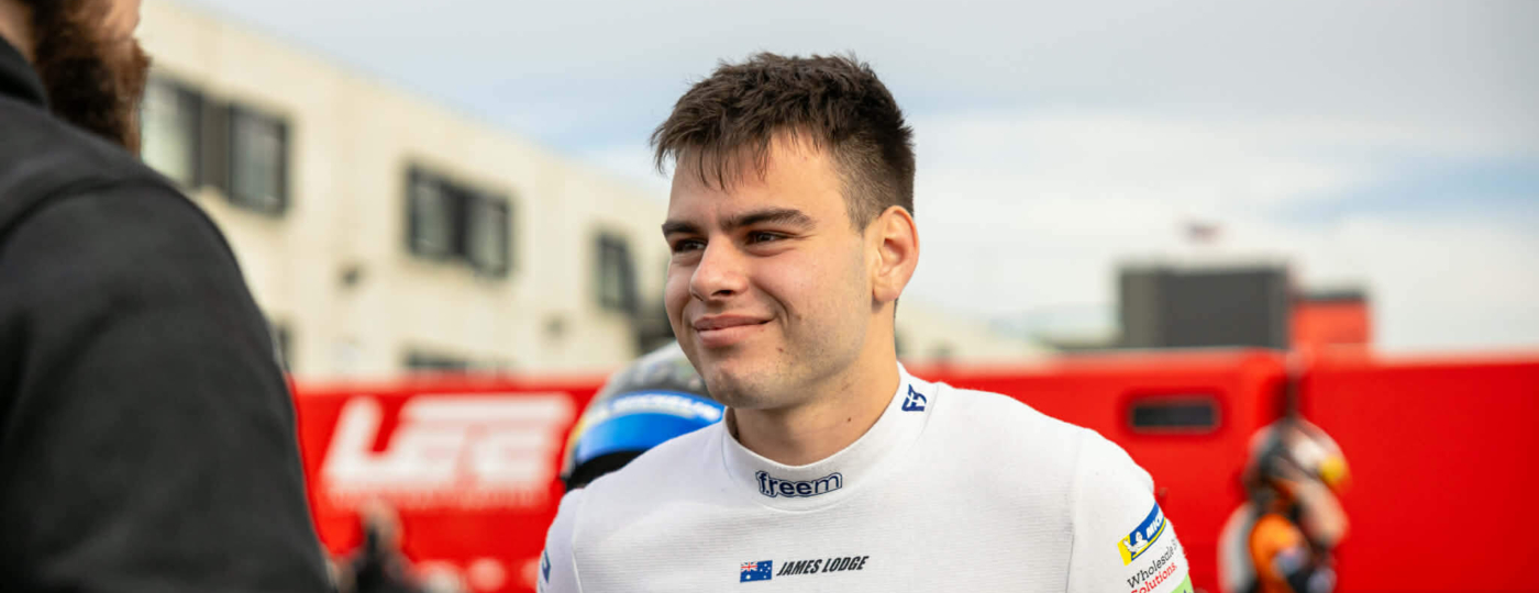 Race car driver James Lodge in a white racing suit greets someone in the pit area with a smile, red barriers in the background.
